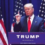 Man speaking at podium with USA flags behind him.