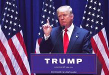 Man speaking at podium with USA flags behind him.