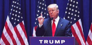 Man speaking at podium with USA flags behind him.