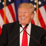 Smiling man speaking at podium with American flags.