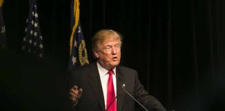 Man speaking at podium with "TRUMP" sign.