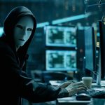 Person wearing mask and hoodie at computer desk.