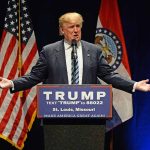 Man speaking at podium with American flags behind.