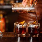 Bartender pouring cocktails into glasses with lemon garnish.