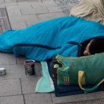 Person sleeping on sidewalk in blue sleeping bag.