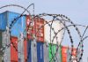 Barbed wire fence with shipping containers in background.