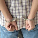 A person in handcuffs viewed from behind wearing a plaid shirt and denim jeans