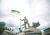 Soldier on tank holding Ukrainian flag.