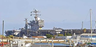 A large naval aircraft carrier docked in a harbor with smaller boats in the foreground