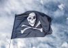 Somali Pirates RESURGE – Special Forces Deployed A black pirate flag featuring a skull and crossbones waving against a cloudy sky