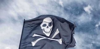 A black pirate flag featuring a skull and crossbones waving against a cloudy sky