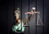 Blindfolded Lady Justice statue holding scales behind bars.