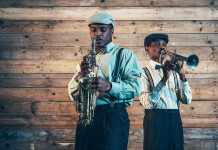 Two jazz musicians playing saxophone and trumpet against a wooden wall