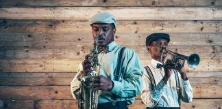 Two jazz musicians playing saxophone and trumpet against a wooden wall