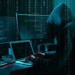 Hacker in dark room surrounded by computer screens.