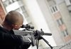 Person aiming a rifle from a building balcony.