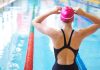 A female swimmer adjusting her pink swim cap at the edge of an indoor swimming pool