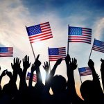 Silhouetted crowd holding American flags against a sunset sky