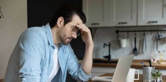 Man stressing over work on laptop at kitchen table.