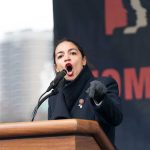 AOC’s Bold Stance Halts Strange Political Alliance Woman speaking passionately at podium during outdoor event.