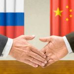 Secretive Ceremony: Russia-China’s Hidden Commitments Two hands shaking in front of two flags.