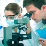 Hollywood Star Sparks Cancer Treatment Frenzy Two scientists conducting an experiment in a laboratory, one looking through a microscope and the other holding a flask with blue liquid