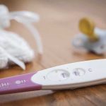 A pregnancy test next to a pacifier and a knitted baby hat on a wooden surface