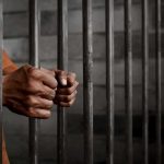 Governor Draws Hard Line On Inmate Care Hands gripping prison cell bars.