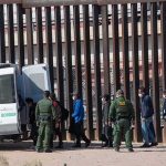 Border agents escort a group near a tall fence and a patrol van