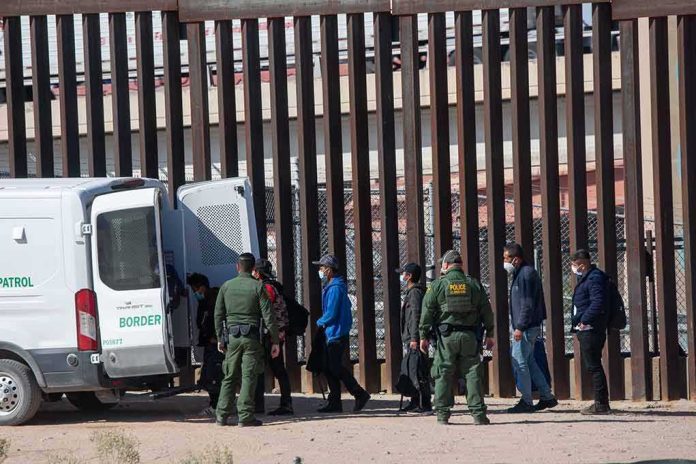 shutterstock_2079408475 Border agents escort a group near a tall fence and a patrol van