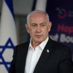 A political leader speaking at a press conference with the Israeli flag in the background