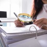The Mass Shooting Number Game Person using a magnifying glass to review documents in a binder