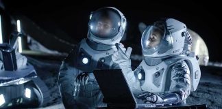 Two astronauts in spacesuits review data on a laptop on a moonlike surface at night