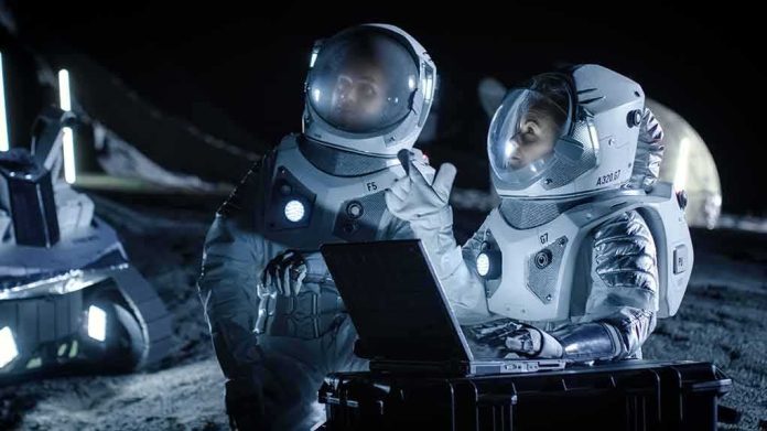 shutterstock_1042736263 Two astronauts in spacesuits review data on a laptop on a moonlike surface at night