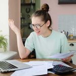 Trillion-Dollar Aid, Misleading Divide Young woman with glasses frustrated at laptop while holding bills