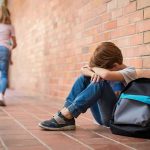 School Gender Switch Kept From Parents? boy sitting against brick wall with head on arms and backpack