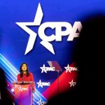 Woman in red speaks at podium on brightly lit conference stage