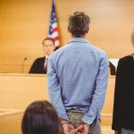 Travis Kelce Named In Shocking Ponzi Fallout Defendant in handcuffs stands before a judge in a courtroom