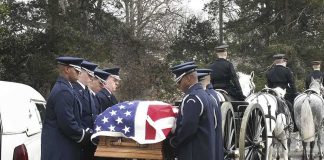 Military honor guard carries a flag-draped casket beside a hearse and caisson