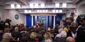 Press Passes Flagged – Chaos At Gates Press secretary at podium addressing reporters in briefing room