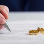 Hand signing a document beside two gold wedding rings