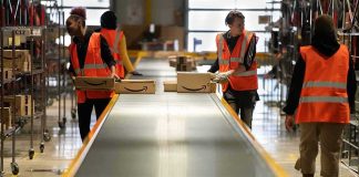 Tesla Firing Shadows Triple Murder Warehouse workers in orange vests handle packages on a conveyor belt